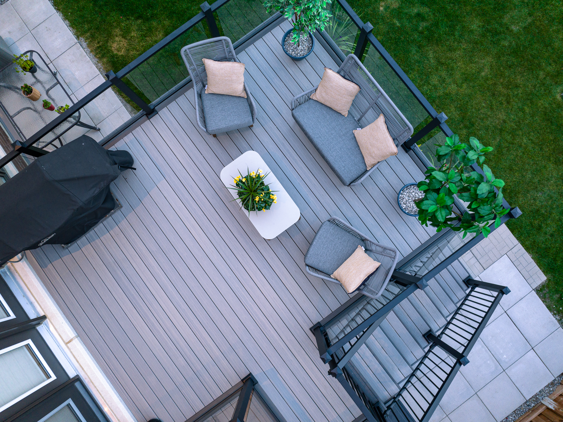 Aerial view of a Fiberon Chai deck with Fortress-Railings1 showcases gray flooring. Features include a seating area with gray chairs and beige cushions, a white coffee table with plants, another decorative plant, and a covered grill. Its all beautifully enclosed by a wooden fence and lush lawn.