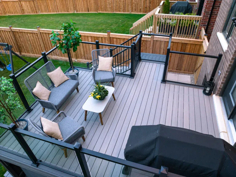 Aerial view of a Fiberon Chai deck with Fortress-Railings1 showcases gray flooring. Features include a seating area with gray chairs and beige cushions, a white coffee table with plants, another decorative plant, and a covered grill. Its all beautifully enclosed by a wooden fence and lush lawn.