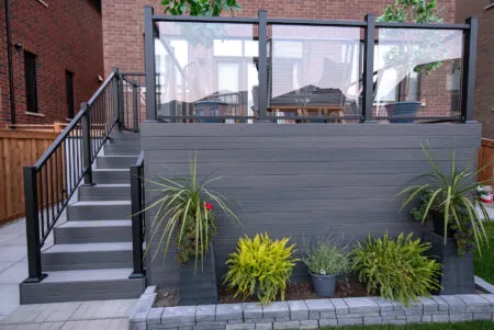 A modern Fiberon Chai deck features gray flooring and a seating area with four gray chairs, each adorned with a beige cushion. A white rectangular table holds a potted plant with yellow flowers. The deck, encompassed by Fortress Railings4 and a wooden fence, offers contemporary charm.