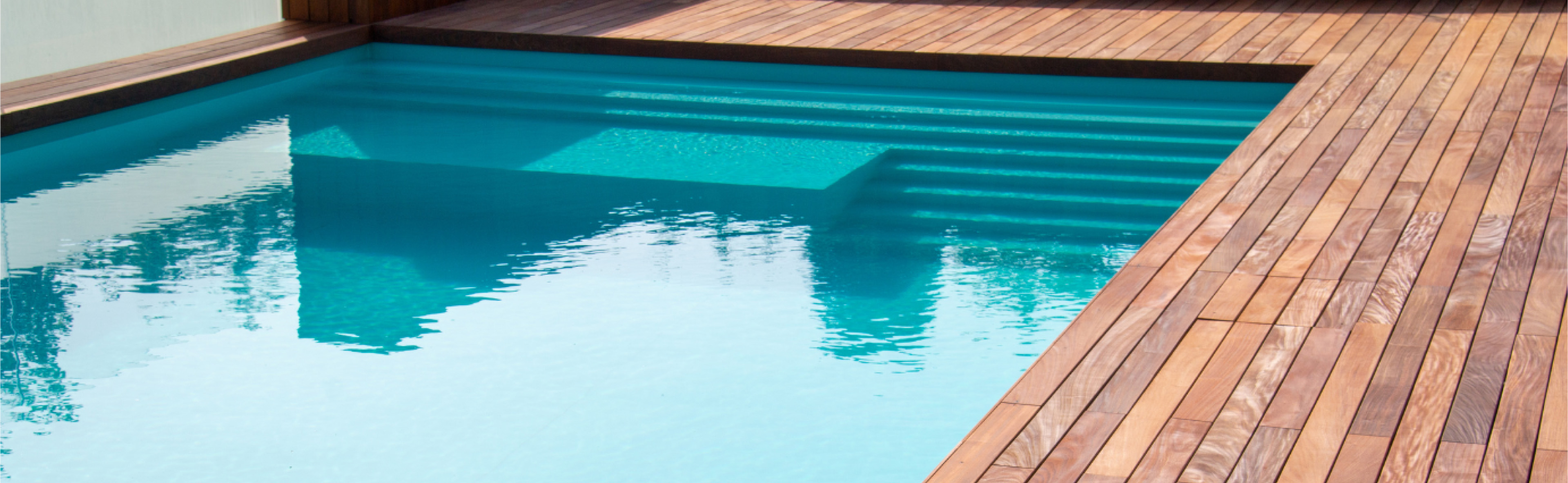 A serene swimming pool with clear blue water bordered by a polished wooden deck. The sunlight casts reflections on the water, and steps lead into the shallow end of the pool.