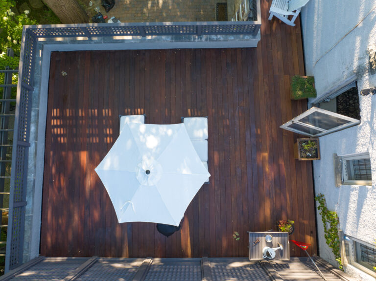 Aerial view of an IPE deck featuring a white patio umbrella and reclining chairs, complemented by additional seating along the perimeter. The area is framed by a gray railing, with lush green trees providing a serene backdrop.