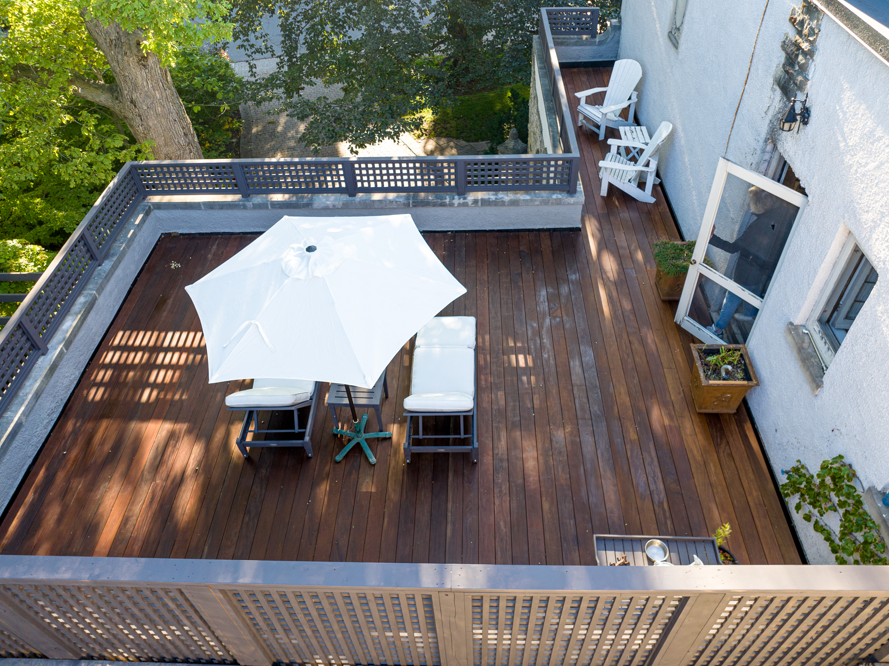 Aerial view of an IPE deck featuring a white patio umbrella and reclining chairs, complemented by additional seating along the perimeter. The area is framed by a gray railing, with lush green trees providing a serene backdrop.