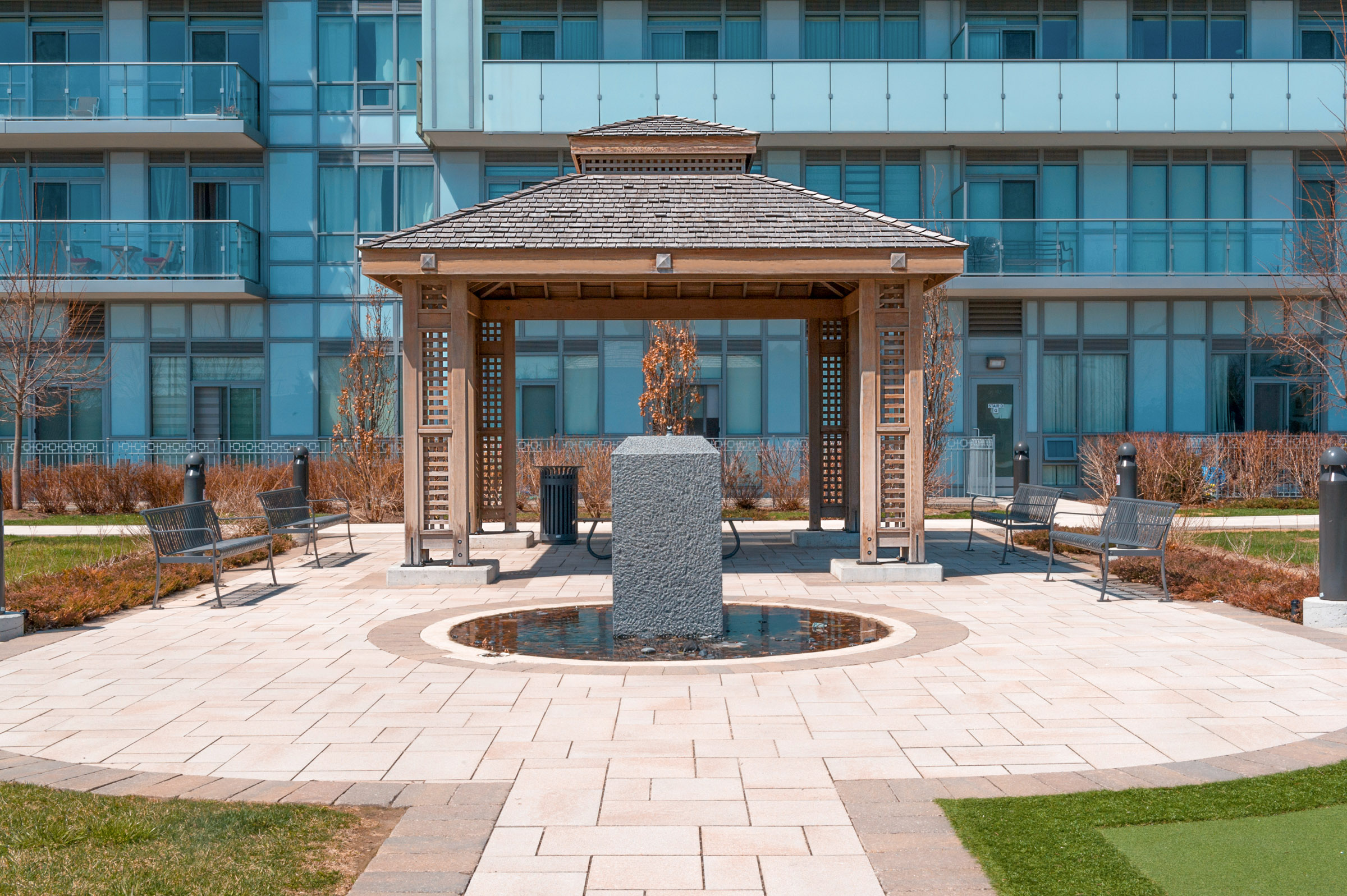 A wooden gazebo with a shingled roof, crafted by Delta Decks Toronto, stands on a paved area in front of a modern glass building. A stone fountain is at the center, surrounded by benches. Green grass and bare shrubs line the walkway.