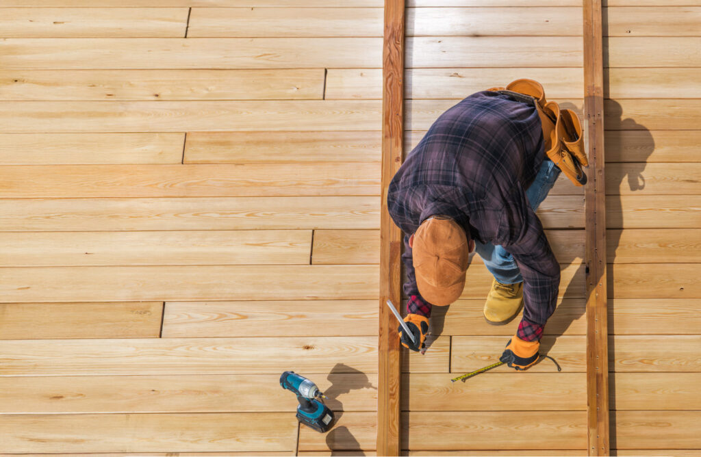 A deck builder at work, wearing a plaid shirt and cap, kneels on a newly installed wooden floor. With precision, he measures with a tape measure amidst wooden beams, a pencil in hand, and a power drill lying close by.