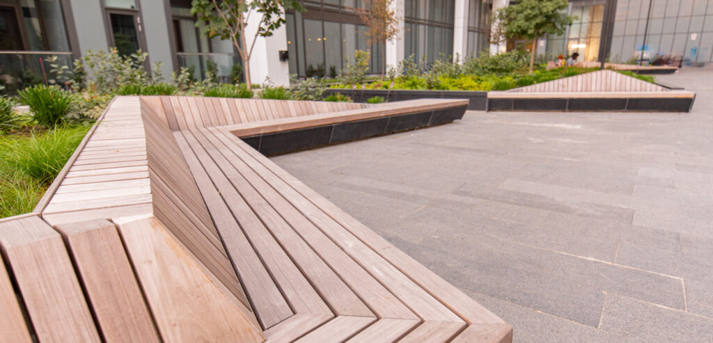 Wooden benches arranged in angular, geometric shapes are set against a contemporary building backdrop. The area features greenery and trees integrated into the design, with large windows and paving stones creating a modern outdoor space.