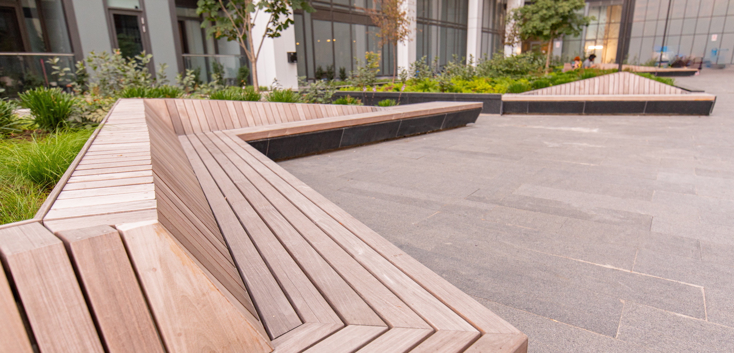 Wooden benches arranged in angular, geometric shapes are set against a contemporary building backdrop. The area features greenery and trees integrated into the design, with large windows and paving stones creating a modern outdoor space.