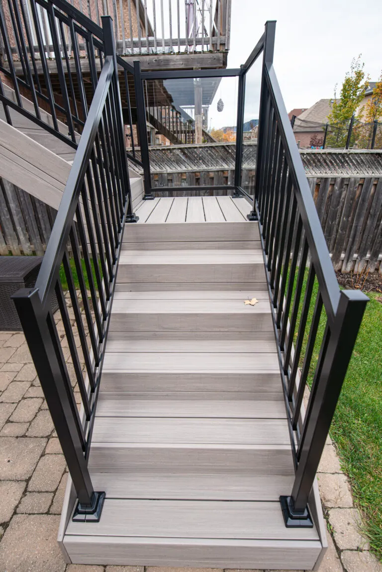 Gray outdoor stairs with black metal railings leading up to a deck. The stairs are surrounded by grass and paving stones, and a wooden fence is visible in the background.