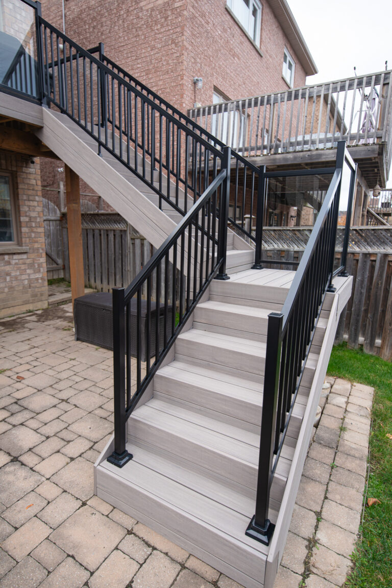 A set of outdoor stairs with light-colored steps and black metal railings, leading up from a paved patio area to a wooden deck attached to a brick house.