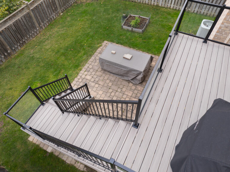A view from above of a backyard showing a raised deck with stairs, black railings, and a patio area with a covered rectangular table on stone pavers, surrounded by a grass lawn and wooden fence.