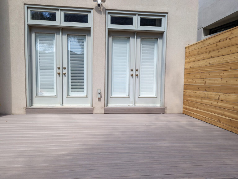 Two white double doors with closed shutters lead to a light gray patio deck. A wooden privacy fence is on the right side, and the wall and surroundings are painted in neutral colors.