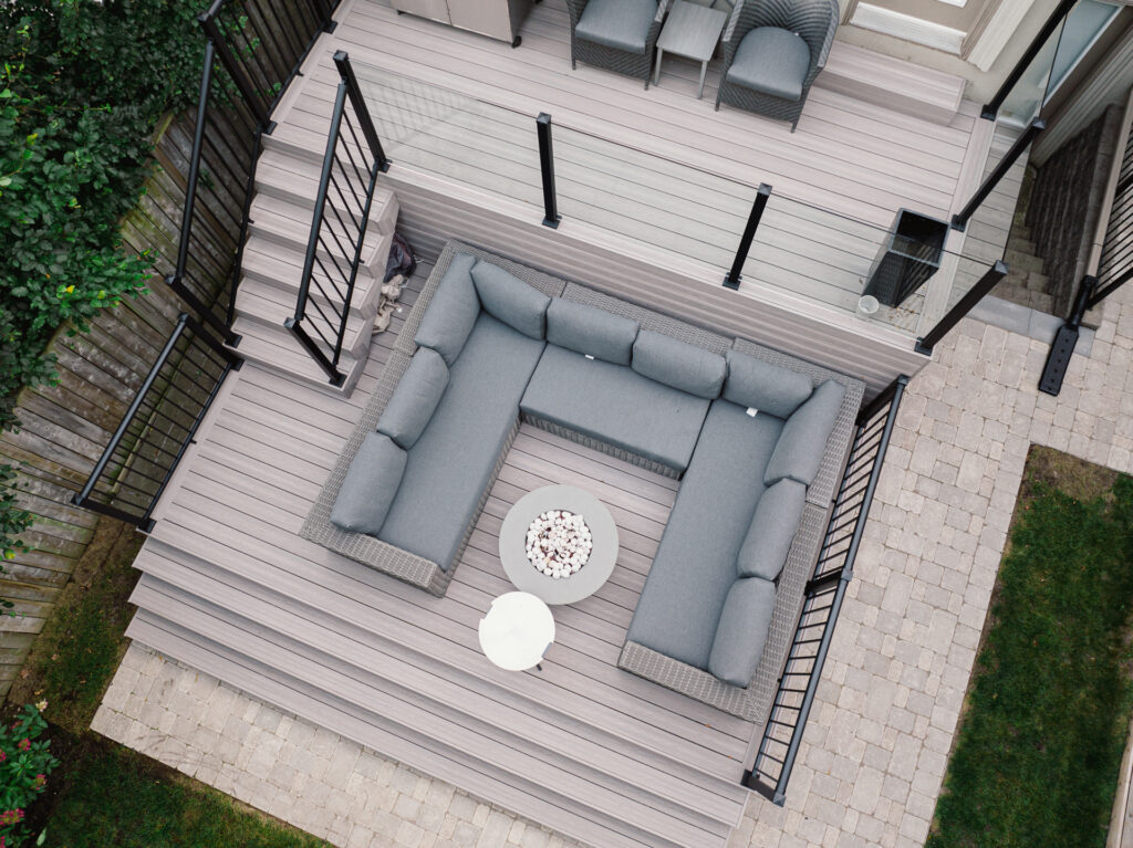 Aerial view of a modern backyard deck with Fiberon Cabana boards, a gray L-shaped sofa, round white table, two armchairs, barbecue grill, and black railings—overlooking a paved patio and lush green grass.