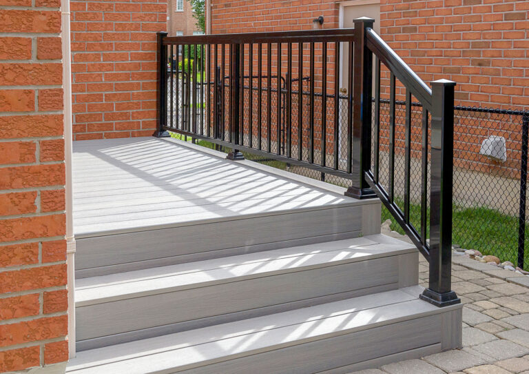 A small set of light gray steps with a matching platform, bordered by a black metal railing, leads up to a brick house with a fenced yard visible in the background.