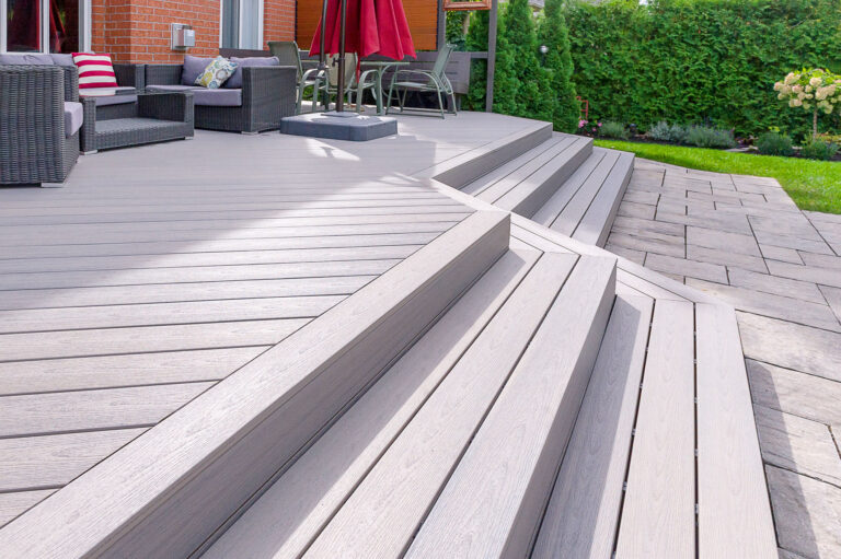 Modern backyard with a multi-level gray composite deck, wide steps, outdoor furniture, a red umbrella, and adjacent patio surrounded by green bushes and grass.