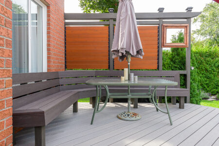 A modern outdoor patio with a gray wooden bench, glass table, and a closed umbrella. The area is enclosed by a privacy screen with wood panels and is surrounded by greenery.