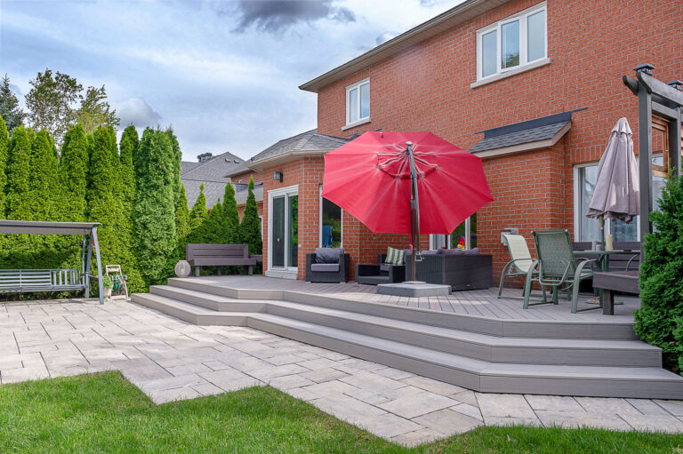 A backyard patio with a raised deck featuring outdoor furniture, a red umbrella, a dining table with chairs, a swing bench, and green shrubs bordering a brick house under a partly cloudy sky.
