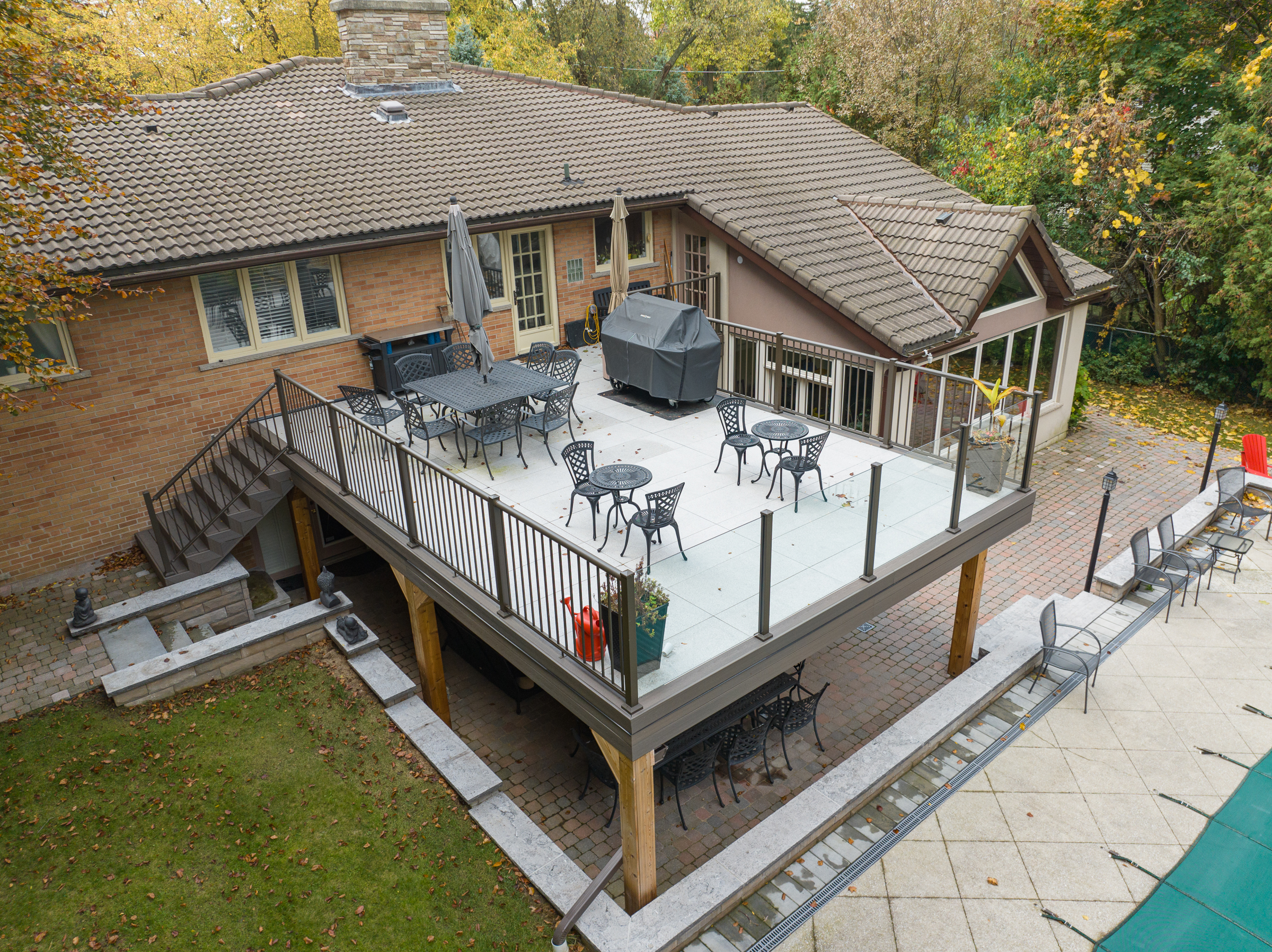A spacious elevated deck attached to a brick house, featuring metal patio furniture, a grill, and umbrellas. Below the deck is a shaded seating area. Surrounding the deck are trees, grass, and a pool area.