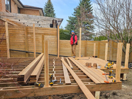 A person in a red jacket stands on a partially constructed wooden deck in a backyard, with tools and wood planks scattered around—a typical scene of Delta Decks at work. Tall wooden fences, trees, and a house frame the background.