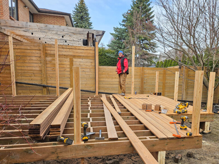 A person in a red jacket stands on a partially constructed wooden deck in a backyard, with tools and wood planks scattered around—a typical scene of Delta Decks at work. Tall wooden fences, trees, and a house frame the background.