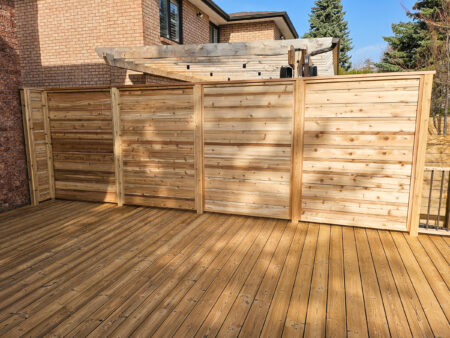 A wooden privacy fence with horizontal slats stands on a wooden deck in a backyard, near a brick house and some trees on a sunny day.