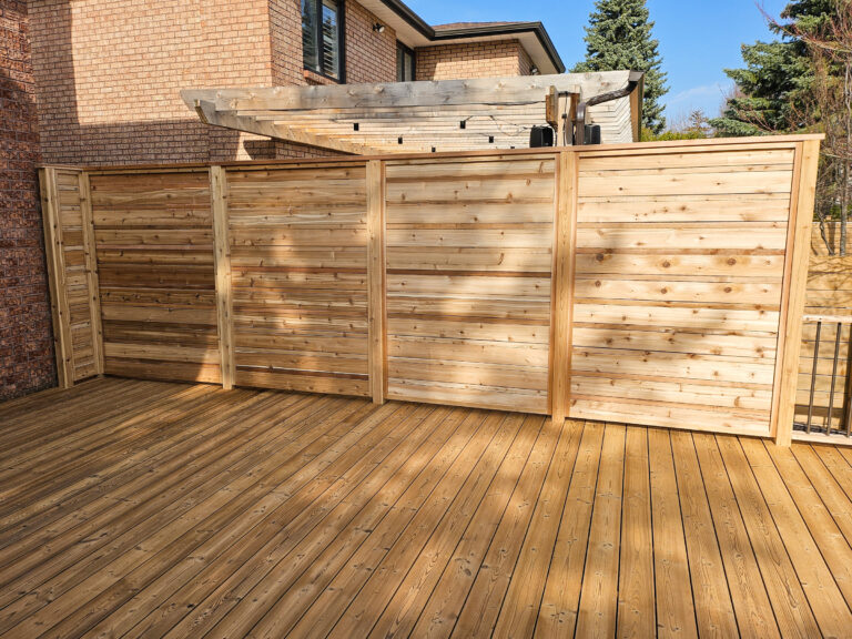 A wooden privacy fence with horizontal slats stands on a wooden deck in a backyard, near a brick house and some trees on a sunny day.