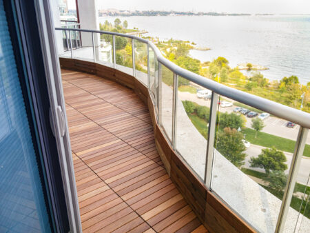 Curved balcony with IPE wooden flooring and glass railing overlooks a park, parking lot, and body of water.