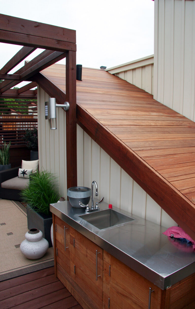 Outdoor kitchen area with stainless steel sink and counter under wooden stairs, adjacent to seating and potted plants.