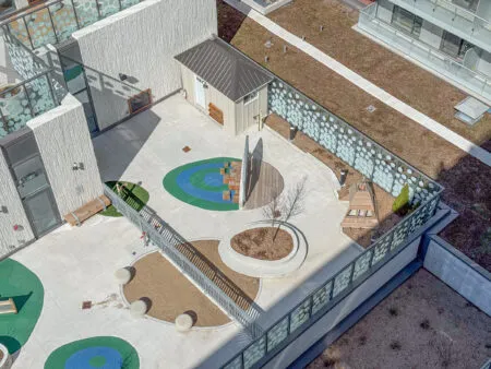 A rooftop courtyard with abstract patterns, seating areas, planters, a shed, and shaded concrete surfaces.