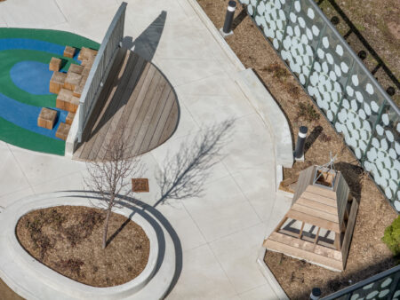 Aerial view of a playground with wooden structures, blue and green rubber flooring, and landscaped areas.