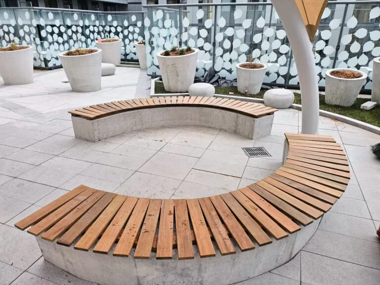 Curved wooden benches with concrete bases in a modern outdoor area with potted plants and a patterned glass railing.