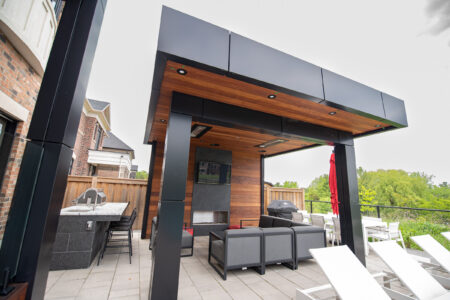 Modern outdoor patio with a wooden ceiling, lounge seating, TV, and built-in grill area next to a fenced yard.