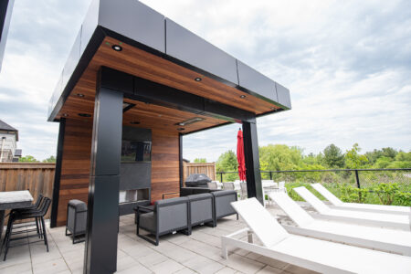 Modern outdoor patio with a wood-paneled pergola, gray seating, lounge chairs, and a red umbrella on a paved surface.