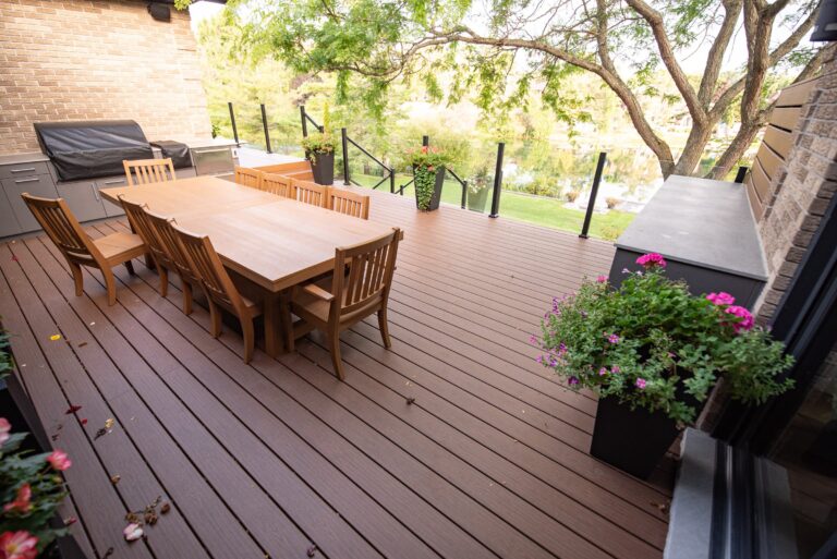 A wooden patio deck with a dining table, chairs, grill, potted plants, and a glass railing overlooking a yard.