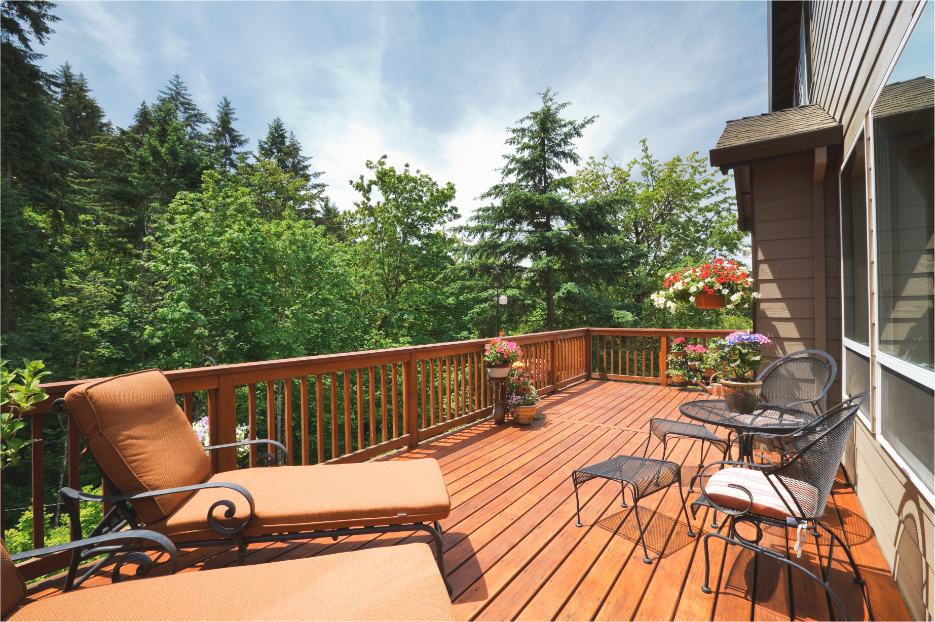 A wooden deck with outdoor furniture, including cushioned lounge chairs and a small table with chairs. Potted flowers add color around the deck. In the background, there is a view of lush green trees under a blue sky.
